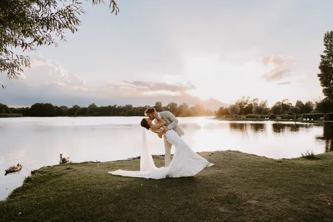 Bride and groom embracing by the lake edge at sunset